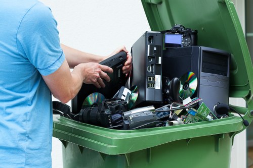 A person wearing a light blue t-shirt is loading discarded electronic devices into a large green wheelie bin, which already contains various broken or unwanted electronic items such as a desktop computer tower, a CRT monitor, a digital camera, and several CDs and electronic accessories. The electronic waste is stacked and spilling slightly from the open lid of the bin, situated outdoors on a paved surface, possibly on a residential driveway or street. The background shows a neutral setting with no additional objects or environmental features visible. The scene reflects rubbish disposal or clearance activities, typical of services offered by offices like Office Clearance Norwood, serving areas around Norwood postcodes. The focus is on the variety of electronic waste materials, emphasizing the importance of proper rubbish management and disposal of electronic items when clearing office or household spaces.