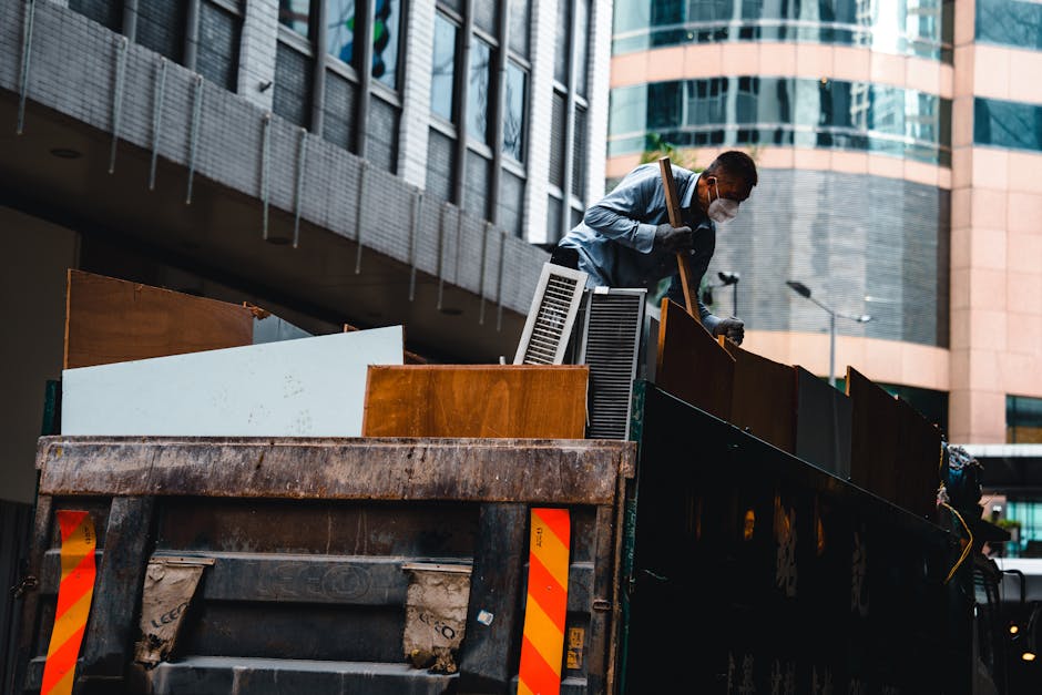 Two men are engaged in rubbish removal outdoors on a clear day, with one wearing a black jacket and yellow gloves, while the other is dressed in a light grey t-shirt. The man in black is holding a car