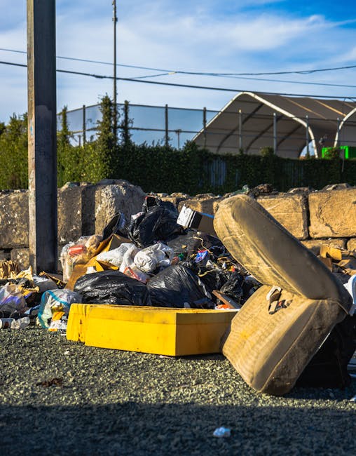Two large, black plastic rubbish bags filled with waste material are positioned on a paved sidewalk in front of a black metal fence and a hedge with dense foliage. The bags appear heavy and are tightl