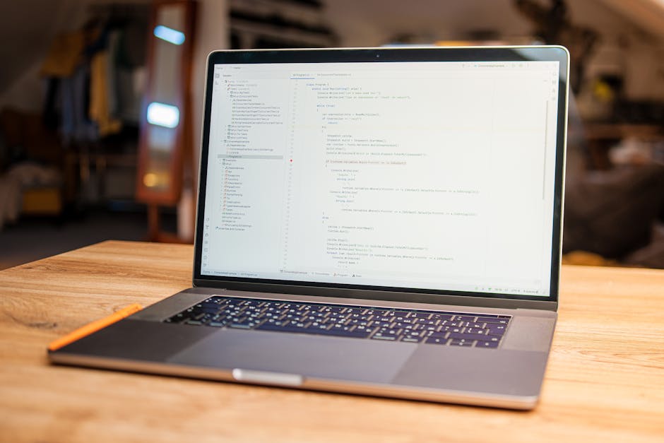 A close-up view of a silver laptop with black keyboard keys placed on a light wooden desk. The laptop screen displays a dark background with lines of colorful programming code. To the left of the lapt