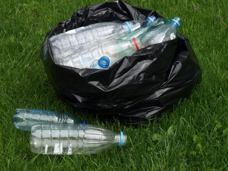 A black plastic rubbish bag resting on a grassy surface, filled with empty clear plastic bottles that have blue caps and are partially visible through the top of the bag. Several bottles, some with red or blue markings, are outside the bag on the grass, indicating they have been recently discarded or removed. The scene appears to be outdoors, possibly in a garden or driveway area, with bright natural lighting. This arrangement reflects typical waste collection for rubbish removal services, such as those offered by Office Clearance Norwood, and illustrates the disposal of plastic beverage containers commonly handled during office or domestic clearance. The focus on plastic bottles highlights the importance of proper waste management and recycling practices for local areas such as Norwood or nearby postcode regions, supporting environmentally conscious rubbish removal solutions.