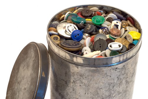 A large, cylindrical metal container with a weathered, slightly tarnished surface, filled to the brim with an assortment of plastic and metal buttons of various sizes, shapes, and colours, including blue, black, white, green, and brown. The container is placed outdoors on a paved surface, with its matching flat lid resting on the ground beside it. The background appears plain and white, emphasizing the focus on the button-filled bin, which is typical of waste collection or disposal in a recycling or rubbish removal context in Norwood or nearby areas. The scene highlights the variety of discarded small household items, and the metal container's utilitarian design supports its use in rubbish collection services such as those provided by Office Clearance Norwood.