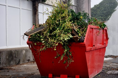 A large, red plastic skip filled with mixed waste, including green foliage, small branches, and wooden pallet fragments, situated outdoors on a paved surface near a white-painted fence and a concrete wall. The skip is positioned on the pavement in a suburban or residential area, with the surrounding environment featuring a wall with climbing plants and a glimpse of trees in the background, indicating an outdoor rubbish clearance setting. The waste materials inside are slightly overflowing, with some greenery hanging over the edge, and the surface of the skip appears textured and weathered, consistent with industrial refuse containers used by office clearance services such as Office Clearance Norwood. The lighting suggests natural daylight, emphasizing the colours and textures of the debris and surroundings, aligning with typical rubbish removal scenes in the Norwood area.