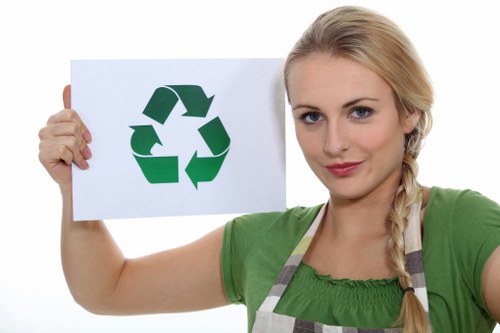 A young woman with long blonde hair tied in a braid, wearing a green top and a striped apron, is holding a white sign with a large green recycling symbol printed on it. She stands indoors against a plain white background, smiling softly at the camera. The recycling symbol consists of three arrows in a continuous loop, indicating recycling and waste management. The woman’s posture is relaxed, with her right arm extended outward to display the sign clearly, emphasizing environmental awareness and responsible rubbish disposal. This image is relevant to office clearance and rubbish removal services offered by Office Clearance Norwood, which operates in the Norwood area with postcode inference around SE27. The setting is neutral, focusing on her and the recycling symbol for clear visual communication in a professional context related to waste management and eco-friendly rubbish disposal in London.